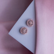 A pair of round embellished rose gold finished silver stud earrings placed on a pale pink surface.